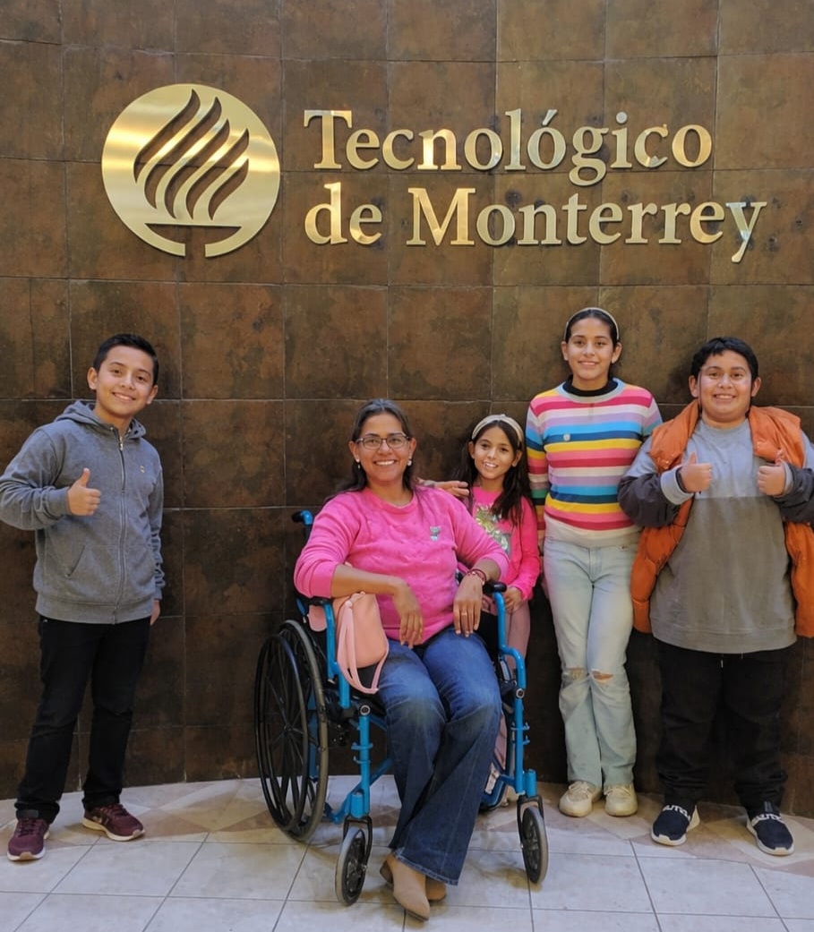 Mujer usuaria de silla de ruedas con niñas y niños frente al logo del Tecnológico de Monterrey.
